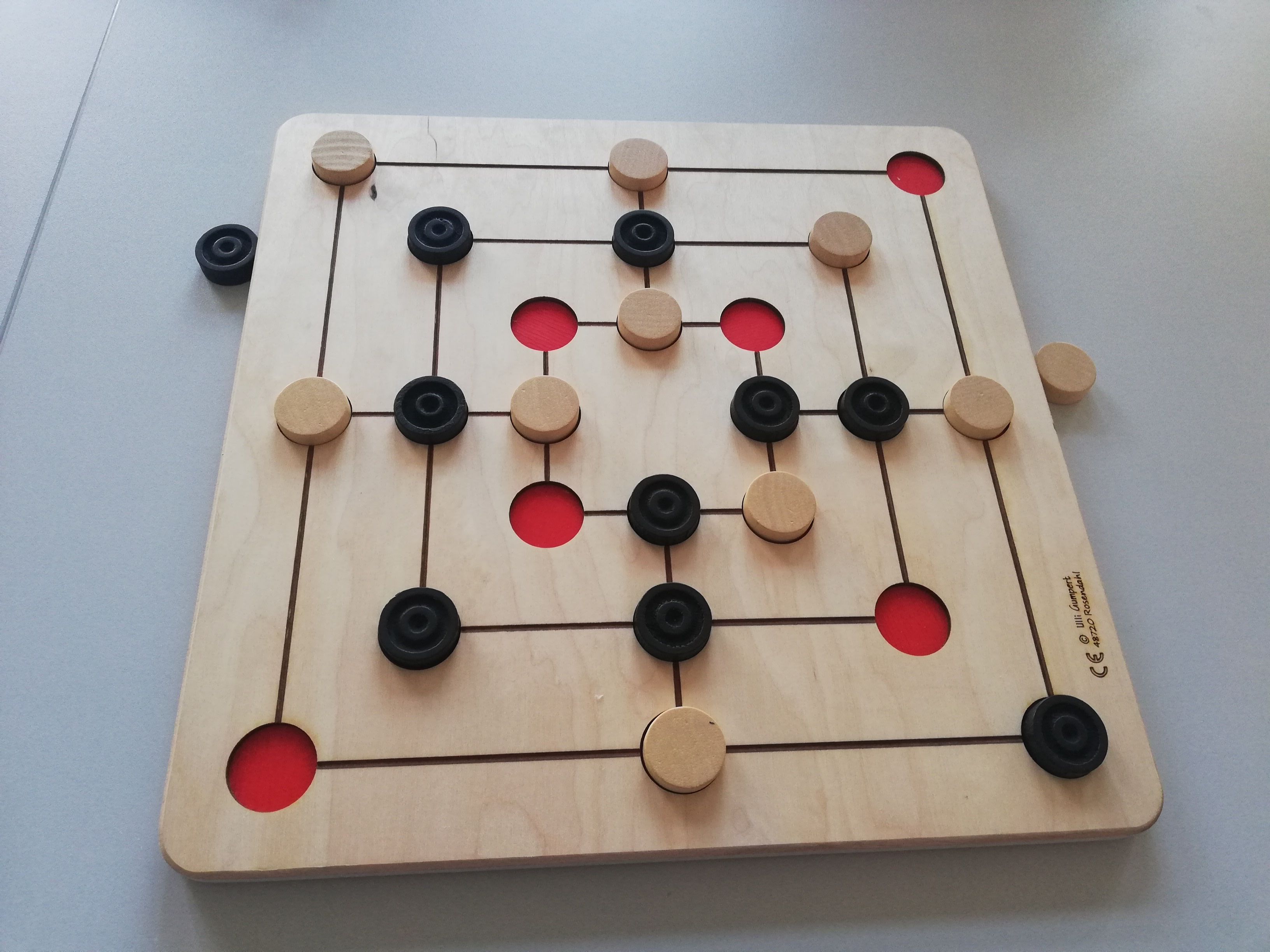 Quadratisches Spielbrett mit ineinanderliegenden Quadraten. Deren Seitenmitten sind über Geraden miteinander verbunden. An den Schnittpunkten und den Ecken sind runde Vertiefungen. Viele Spielsteine in weiß und schwarz sind gesetzt.