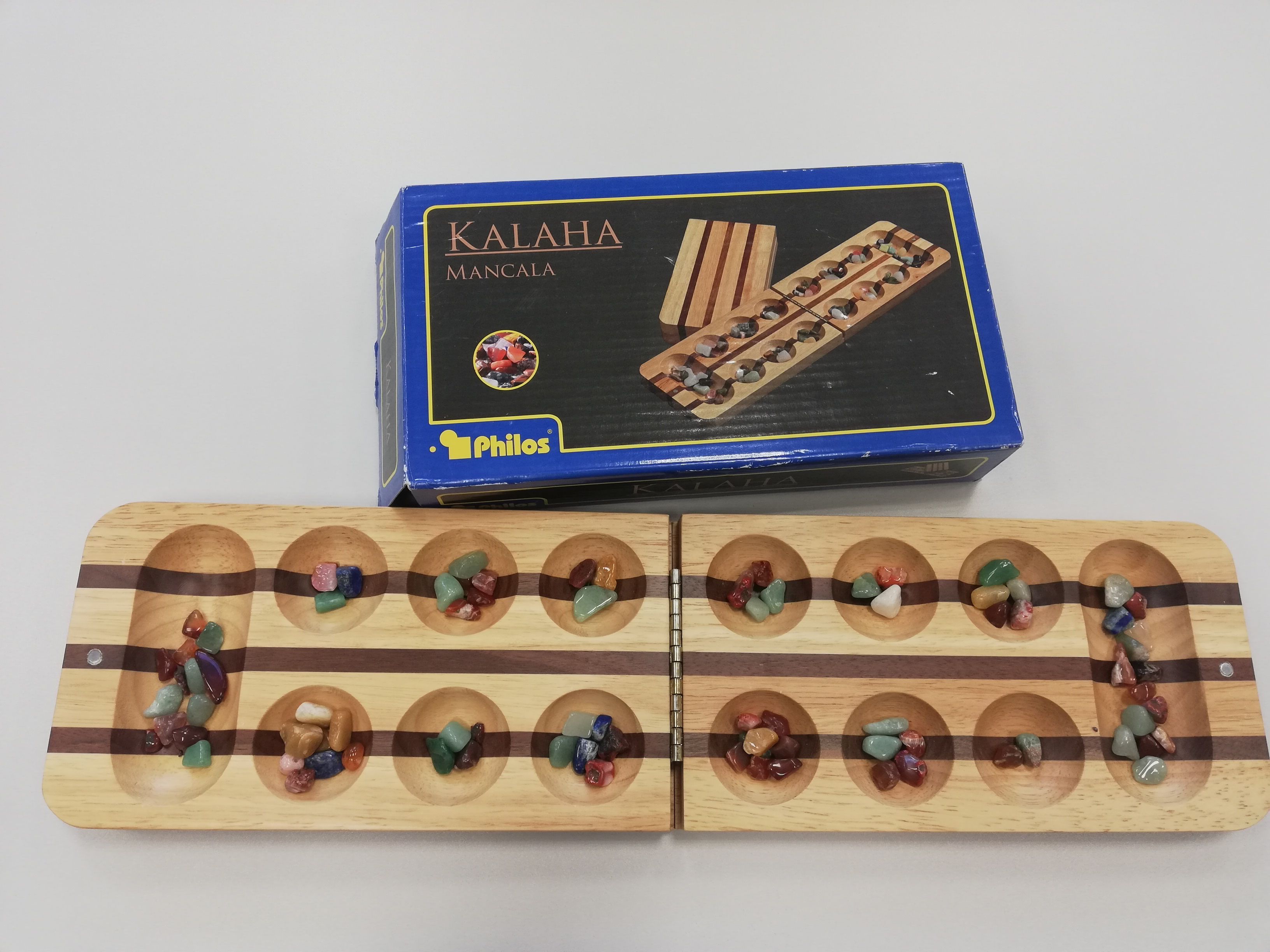 Das aufgeklappte rechteckige Spielbrett hat links und rechts eine Kalaha und dazwischen zwei Reihen runde Mulden. In den Kalaha’s und den Mulden liegen Spielsteine in unterschiedlicher Anzahl.