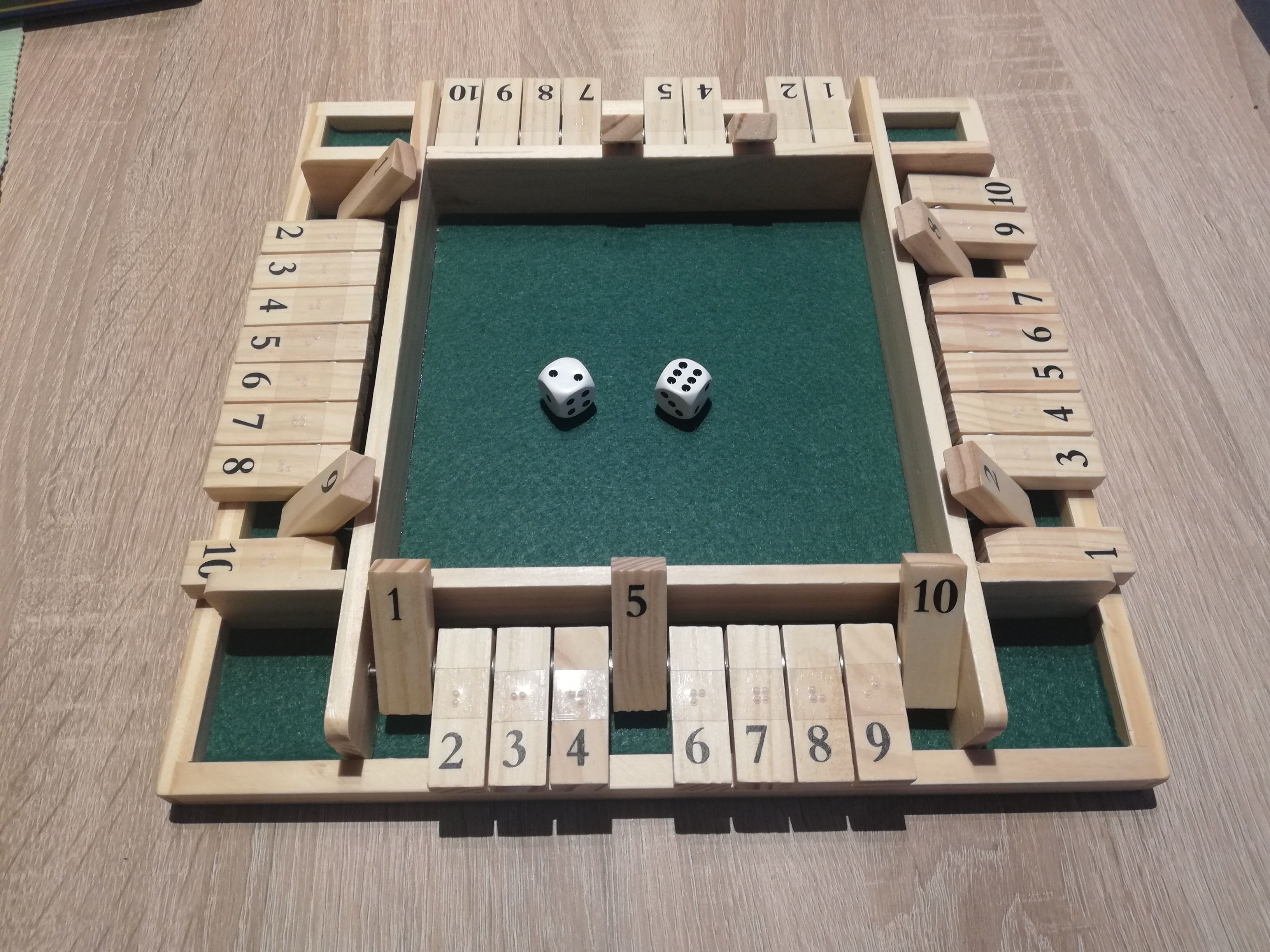 Quadratisches Klappenspiel für 4 Personen. An allen Seiten sind einzelne Klappen, mit Schwarz- und Blindenschrift, umgelegt. In der Mitte auf grünem Grund liegen zwei weiße Würfel mit schwarzen Punkten.