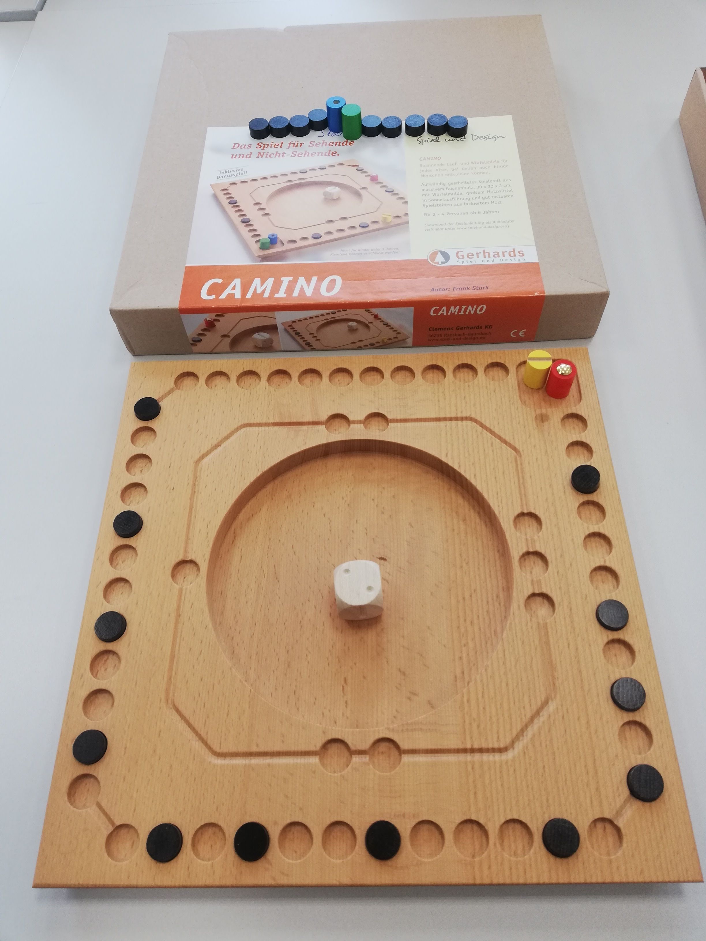 Quadratisches Spielbrett mit einem inneren und äußeren Spielfeld. Eine rote und gelbe Spielfigur stehen im Start- und Zielfeld. 16 Camini's sind auf drei Seiten verteilt. Ein weißer taktiler Würfel liegt in der Mitte des Spielbretts.