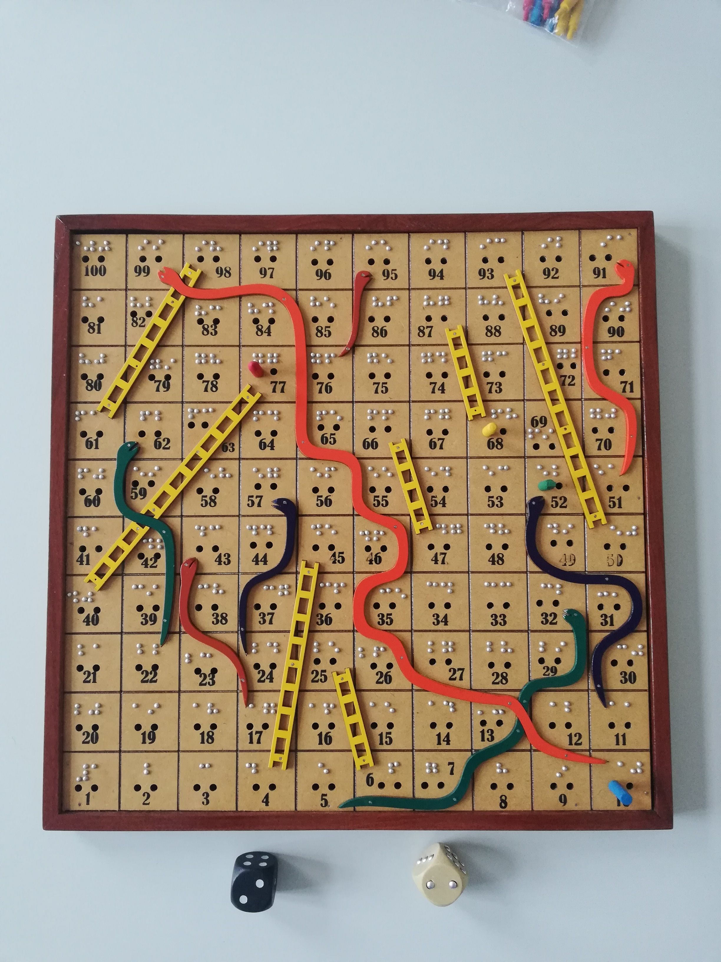 Taktiles quadratisches Spielbrett mit Leitern und Schlangen, Die Spielfelder sind in Blinden- und Schwarzschrift beschriftet. Zwei taktile Würfel liegen davor.